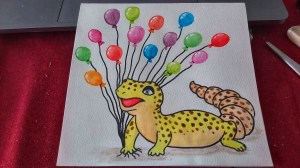 A finished card of a watercolour gecko holding balloons