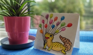 A watercolour gecko card on a windowsill next to a flower