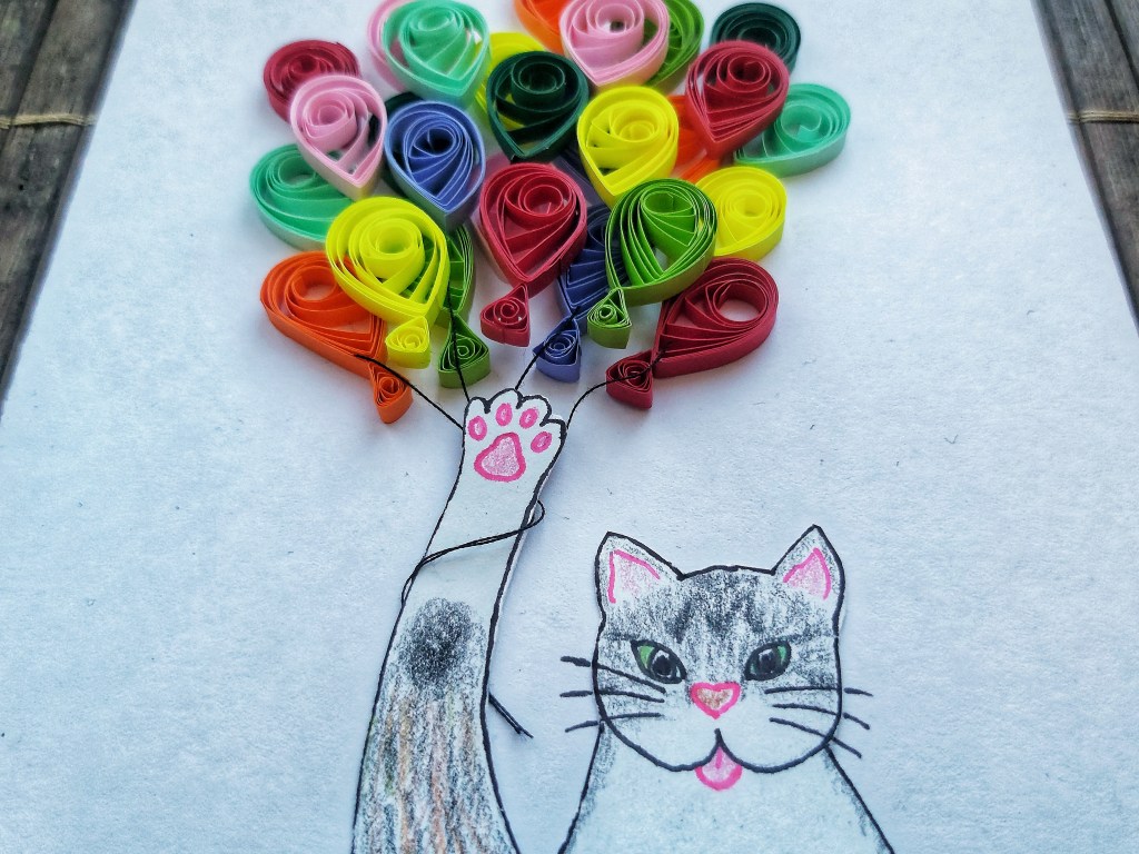 Close up of a handmade greeting card. Face of a cat drawn with coloured pencils. The cat is holding a bunch of quilled colourful balloons in its paw.