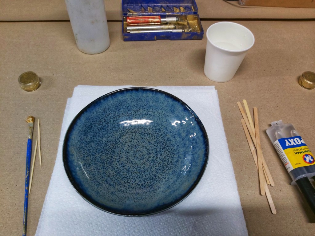 A navy blue ceramic bowl, a syringe with epoxy, a small container of gold powder, a brush and wooden stirrers.