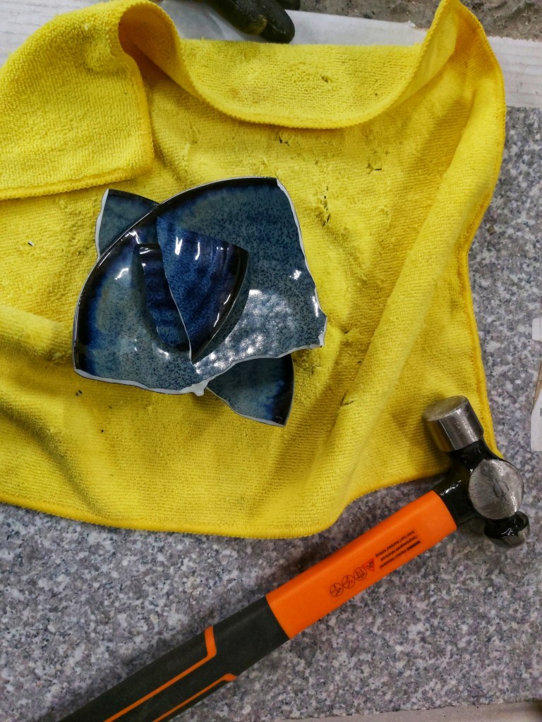 A navy blue ceramic bowl broken into four pieces. Placed on a yellow cloth, next to a hammer.
