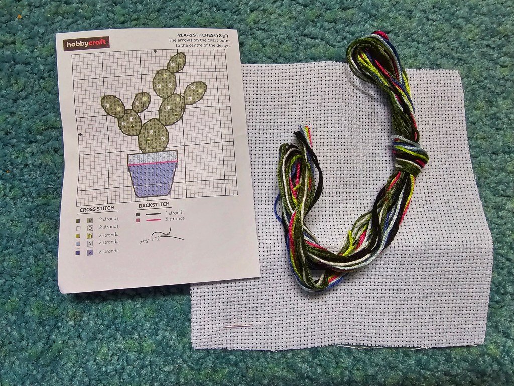 Unpacked mini cross stitch kit containing an aida, a needle, assorted threads and a cross stitch diagram with instructions for making a cactus.