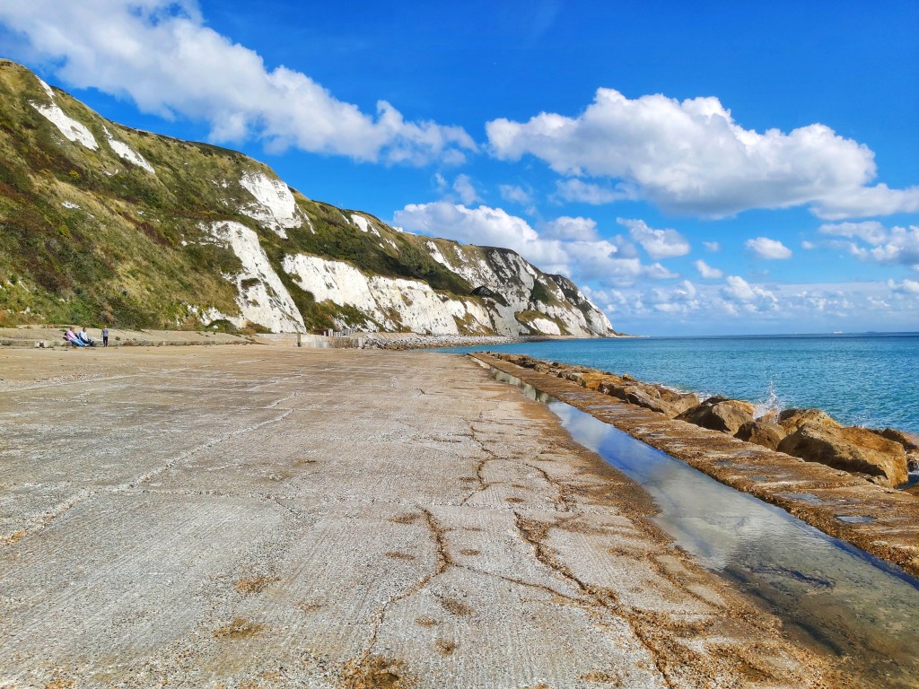 Wide concrete path on the coastline between Folkestone and Dover. Blue sea on the right, while cliffs on the left.