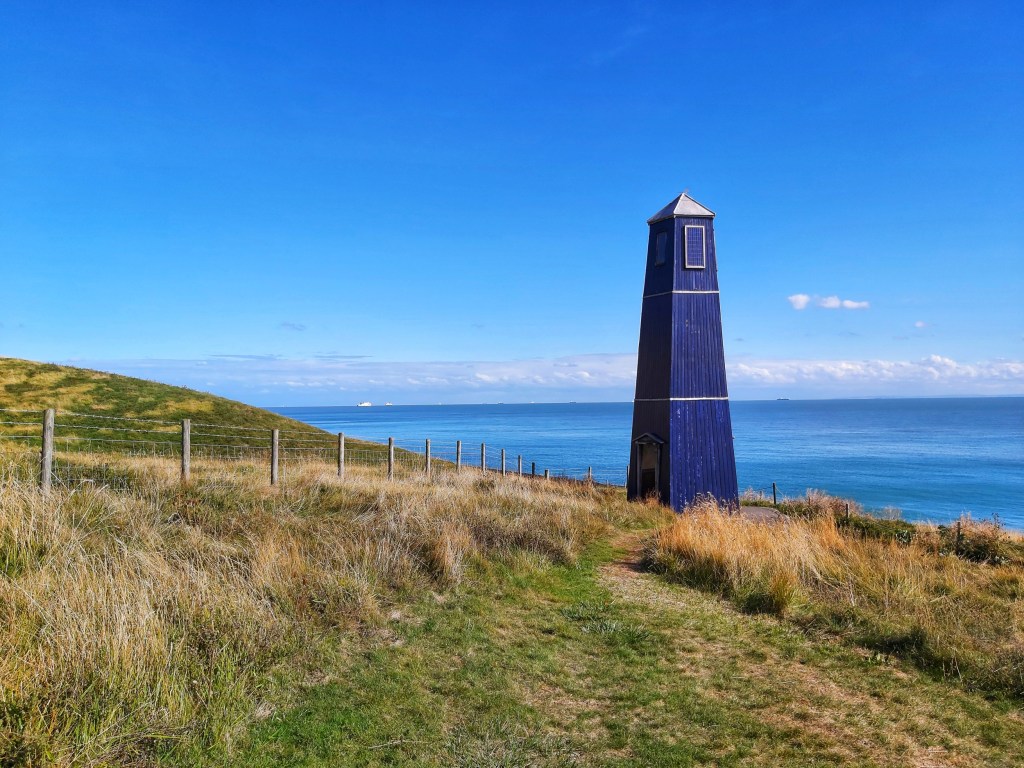 An old, blue, wooden lighthouse between Folkestone and Dover. Sunburnt grass around it. Blue sea on the horizon behind it.