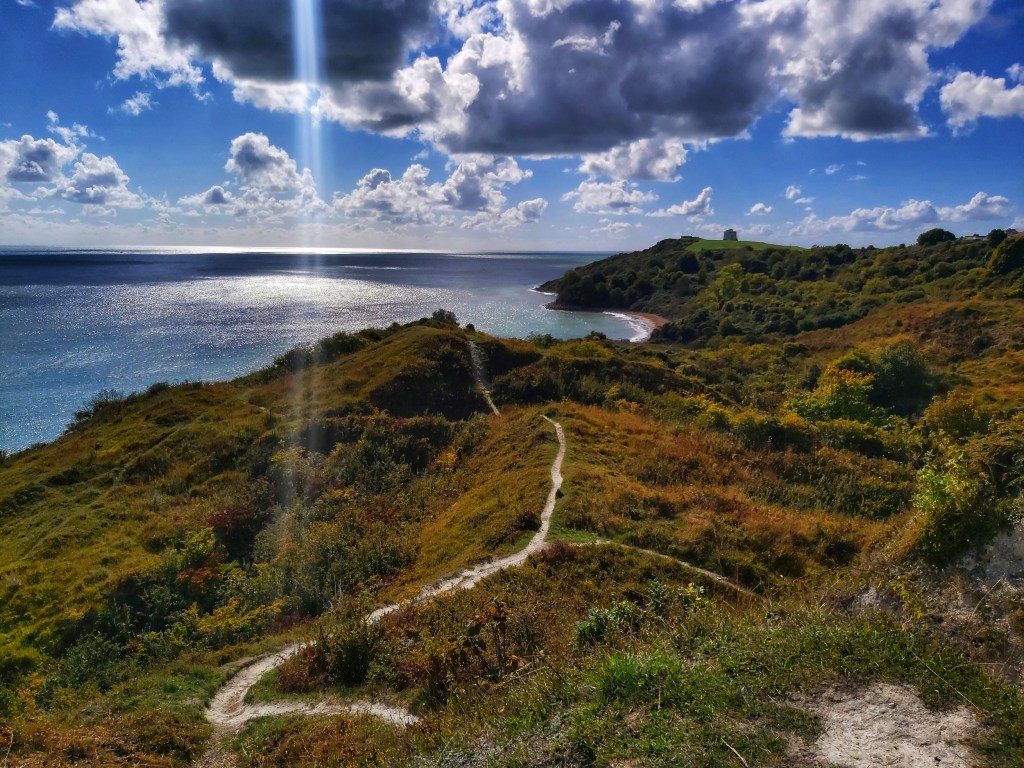 A breathtaking panorama from the top of a cliff on the trail between Folkestone and Dover. Brown, autumnal grasses covering the hills. Blue waters of the English Channel.