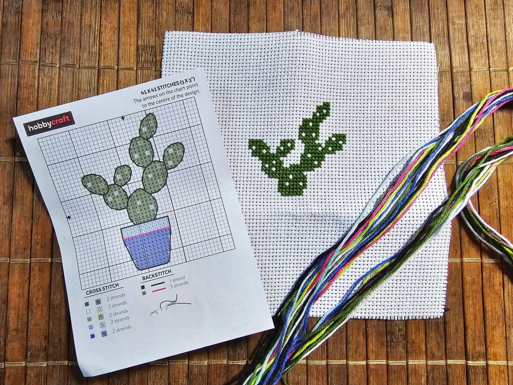 White aida with green cactus leaves cross stitched on. A piece of paper with the cross stitch cactus diagram and a bunch of colourful yarn.
