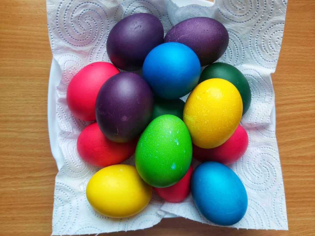 A pile of coloured hard boiled eggs on a paper towel. The eggs' shells were painted in yellow, pink, green, blue and purple.