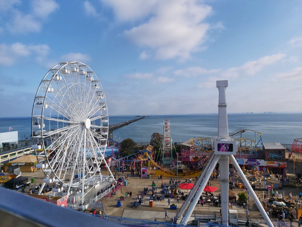 Fun fair on the beach in Southend-on-Sea. View from a hill. Blue skies with a few fluffy clouds. Sea and long Southend pier visible on the horizon.