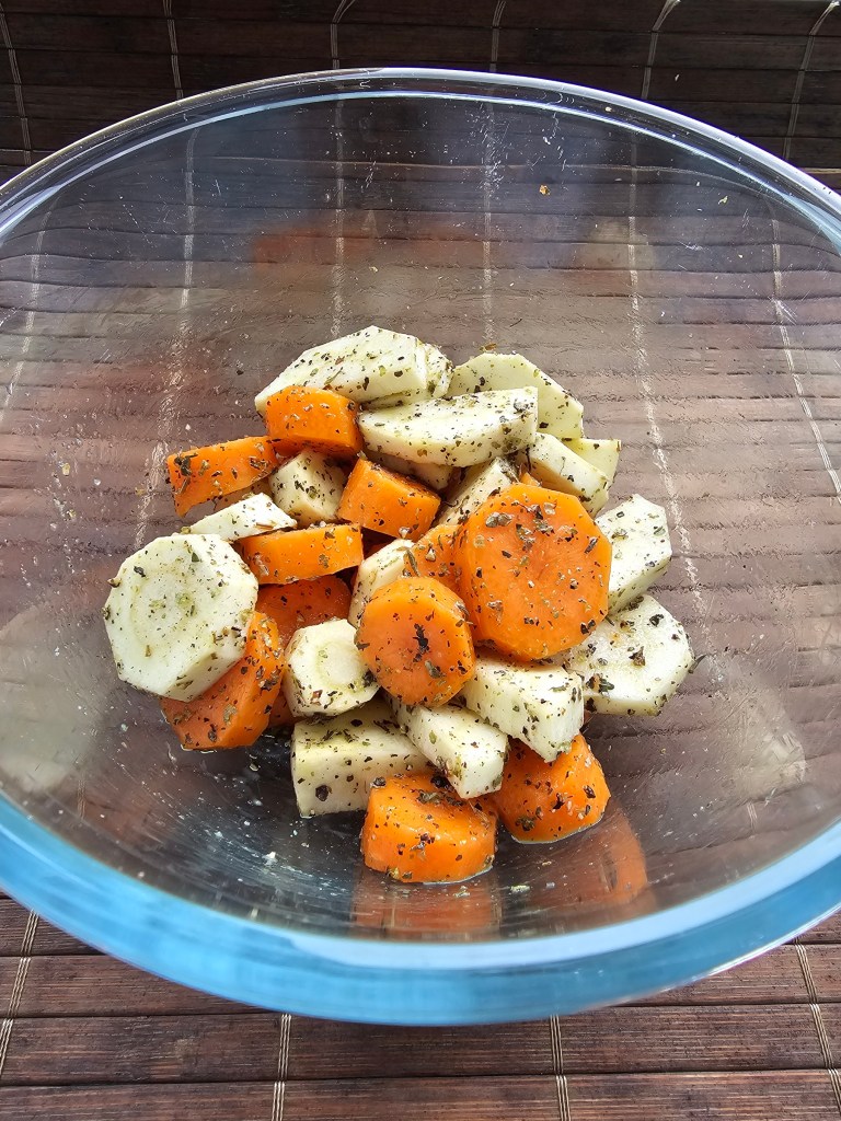 Sliced carrots and parsnips in a glass bowl, covered with olive oil and herbs.