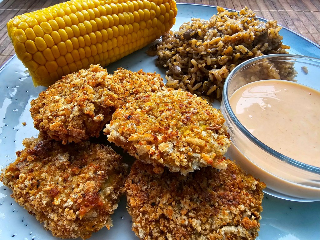 A pile of air fried chicken nuggets on a plate next to a small dish with dipping sauce, a cob of sweetcorn and a pile of rice with mushrooms.