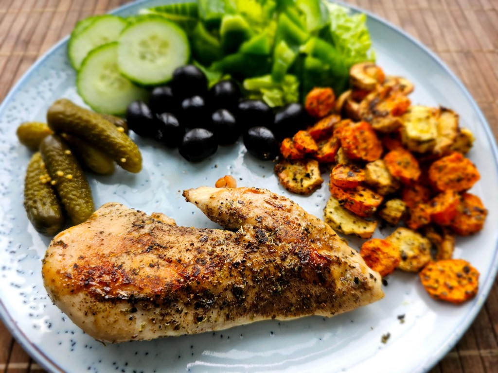 Air fryer roasted chicken breast on a blue dinner plate next to roasted root vegetables, olives, cucumber, lettuce and pickles.
