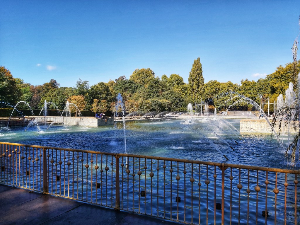 Large fountain in Battersea Park, London. Green trees in the distance.