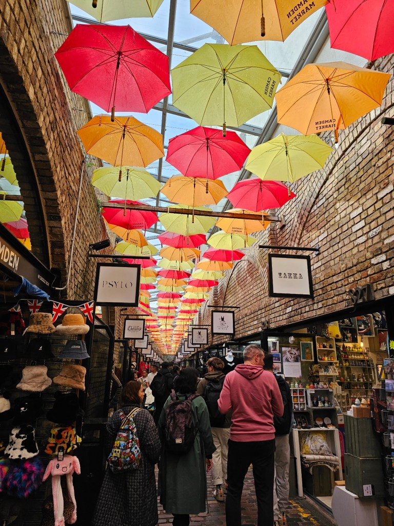 An alley in Camden Market, London, with yellow, orange and red umbrellas hanging under the ceiling. An array of quirky shops on both sides of the alley.