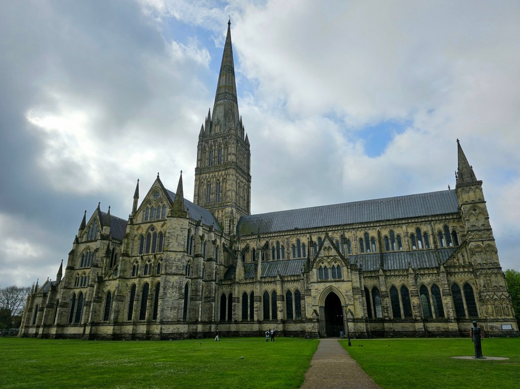 Salisbury Cathedral. 12th century Gothic building with a tall spire. Green, trimmed lawn in front of the cathedral. A little bit of blue skies visible between heavy clouds.