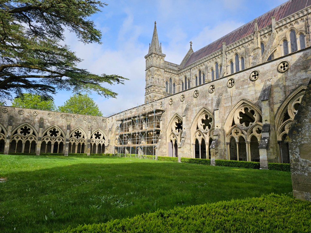 Cloisters of Salisbury Cathedral. Green square patio surrounded by arcades with arched, glass-free windows. Blue skies with light clouds.