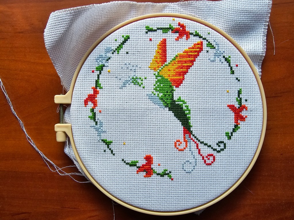Partially completed hummingbird design of a cross stitch kit. Blue, green, red, orange and yellow hummingbird inside a flower wreath.