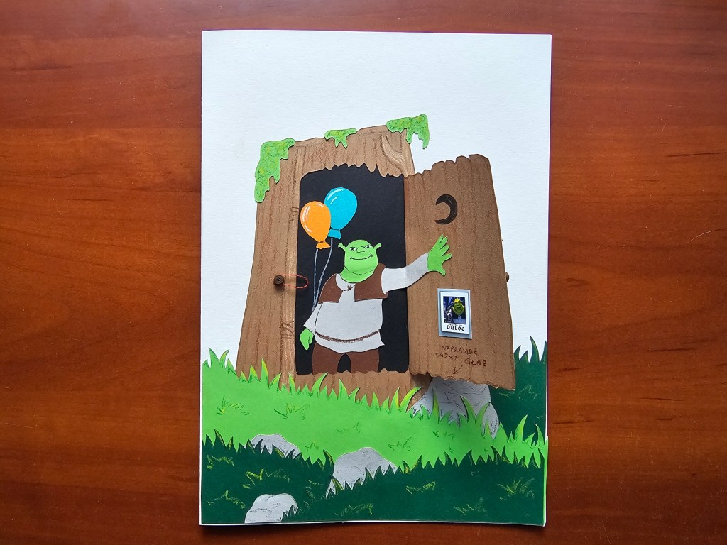 Completed DIY card for a Shrek lover. There's a Shrek figure inside his toilet shed. The shed's door is open. Shrek is holding an orange and blue balloon. There's a photo of Shrek and Donkey hanging on the door.