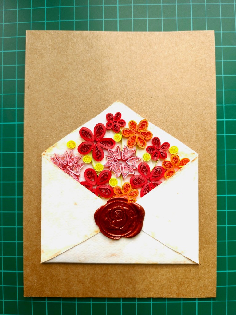 Envelope shape cut out of watercolour paper, folded. The paper was lightly painted with yellow-brown watercolours to make it resemble old parchment. The envelope has a wax seal with a flower design. Inside the envelope, there are quilled pink, red and orange flowers. The whole design is placed on a brown card blank.