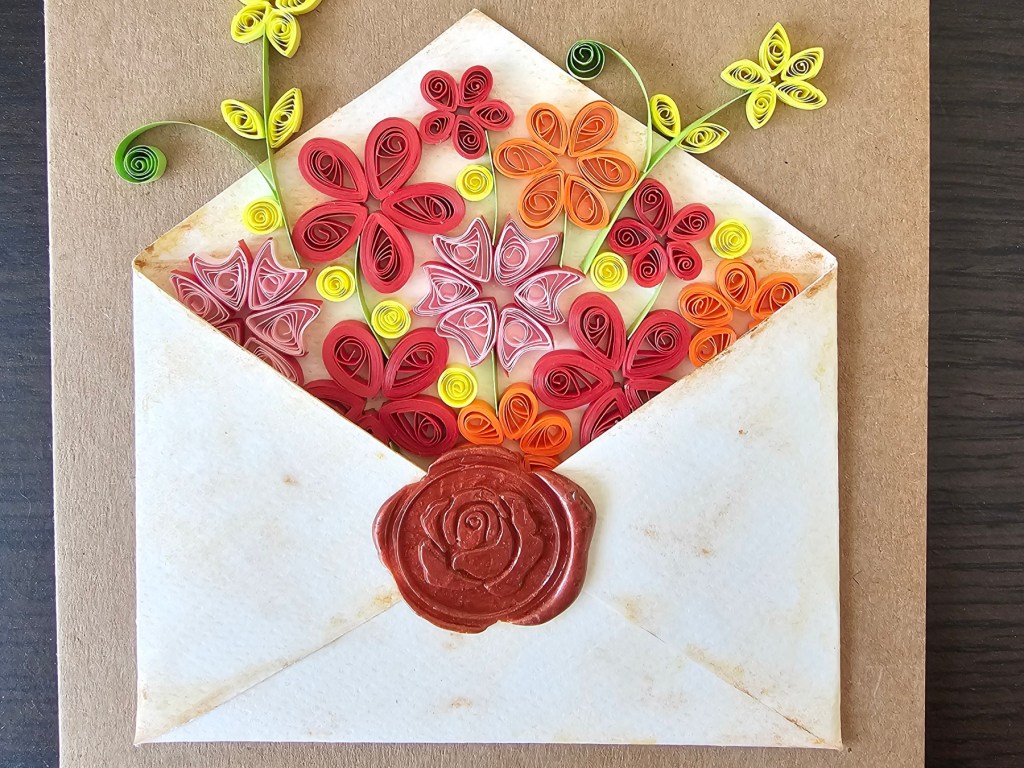 Completed DIY card with an envelope filled with red, pink, orange and yellow quilled flowers. The envelope is placed on a brown card blank.
