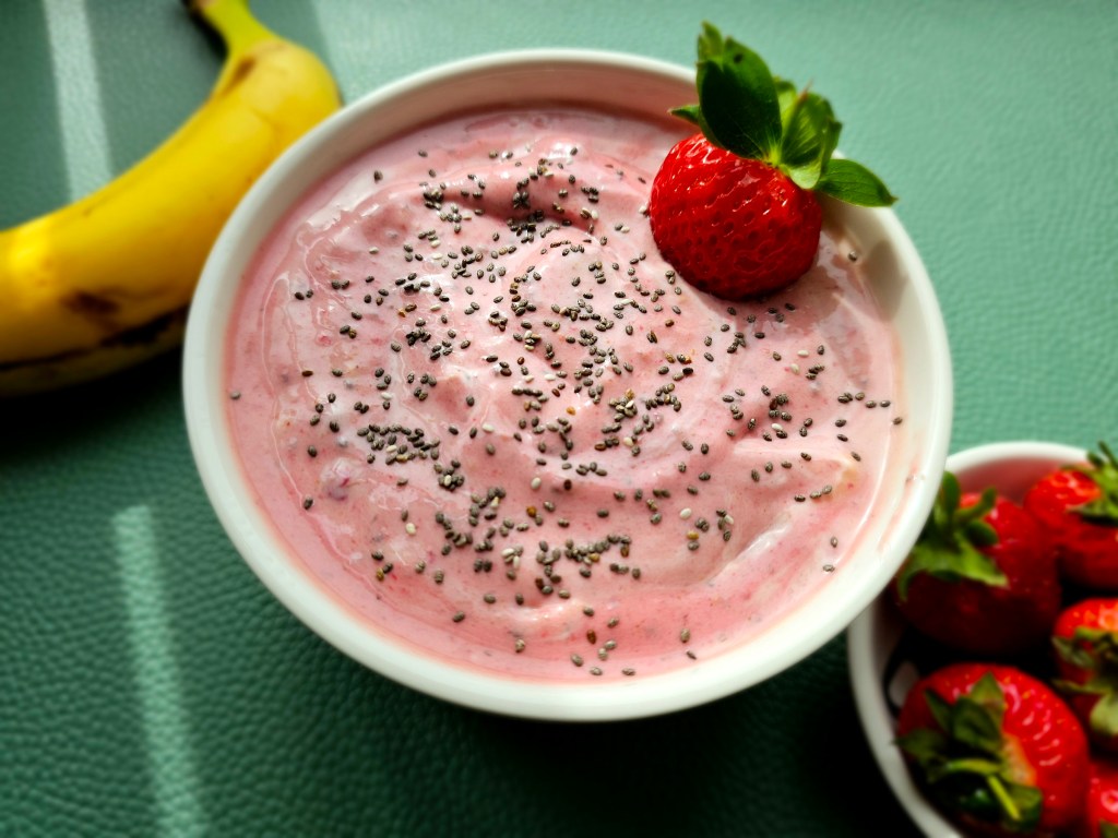 A bowl with frozen smoothie in pink colour. The smoothie is sprinkled with chia seeds and garnished with a fresh strawberry. There's a banana next to the bowl and a small plate with strawberries.