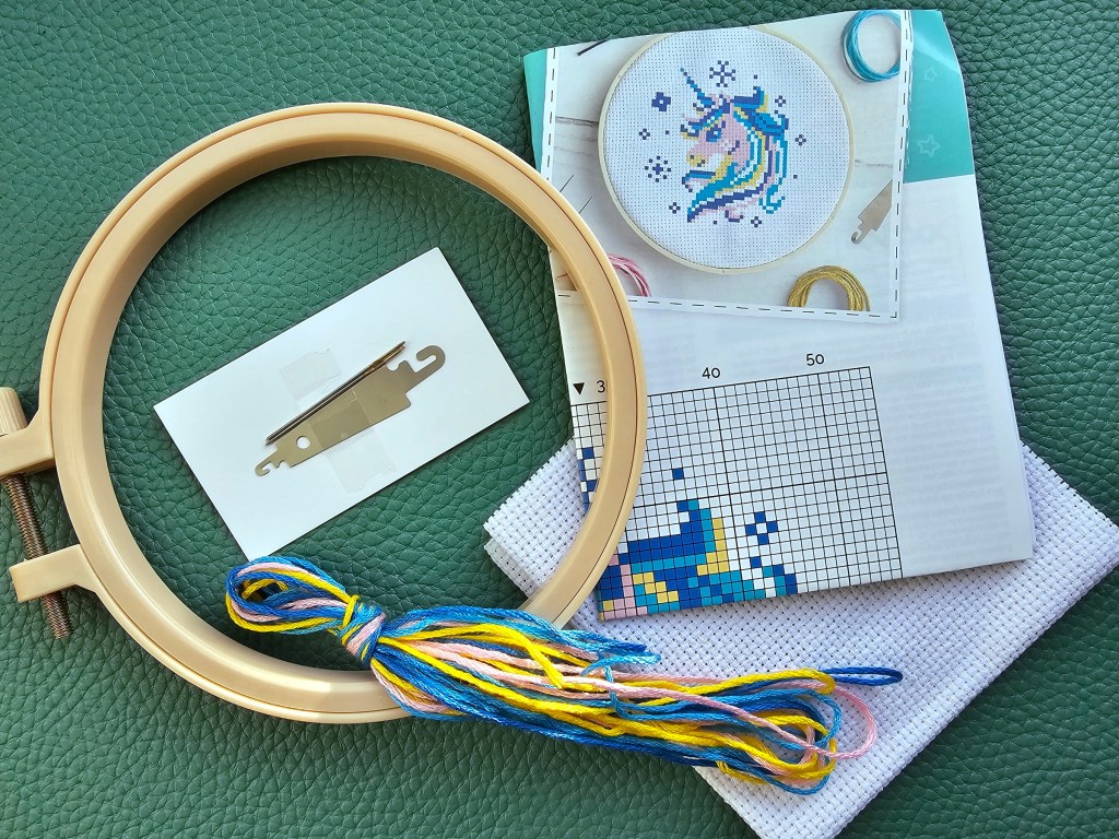 Contents of a mini cross stitch kit with a unicorn design. The kit contains a plastic hoop, assorted threads, needles, threader, blank aida and a leaflet.
