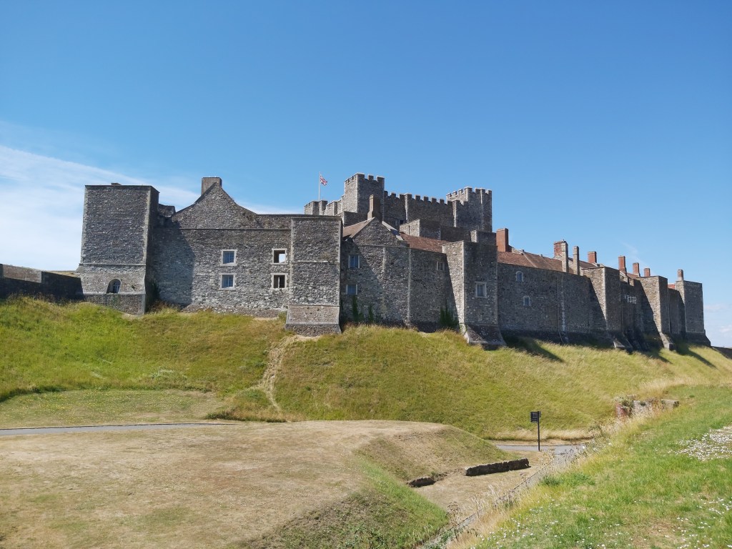 Dover castle viewed from the castle's premises. The castle is located on top of a small grass-covered hill. Blue skies. There's a Union Jack flag on top of the castle.