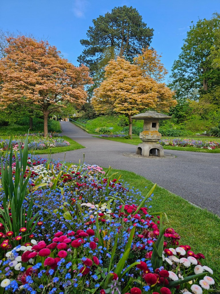 Japanese garden in Folkestone, England. A small, stone decorative house in a Japanese style. Two golden-orange trees behind it. There are also flower beds with a variety of blue and pink flowers.