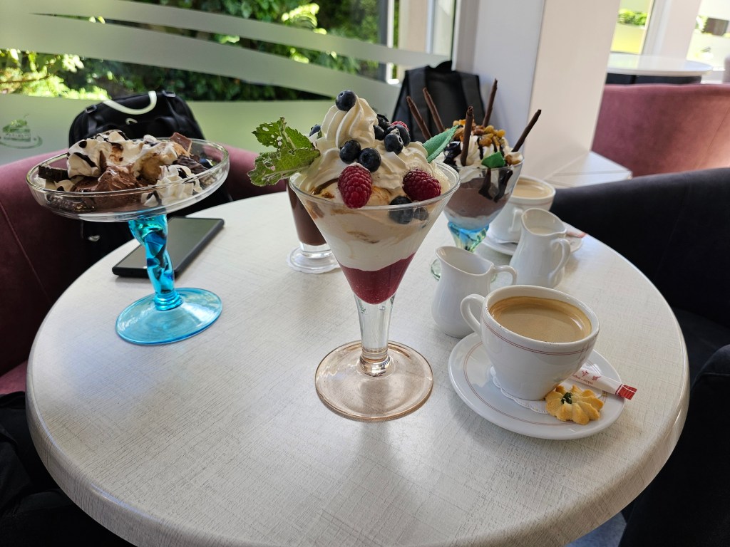 Three large servings of ice cream with whipped cream and other toppings on a table, next to three cups of coffee.