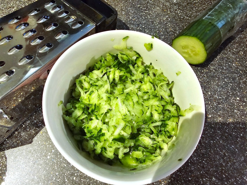 A bowl with grated cucumber shreds, next to a grater and half of a cucumber.