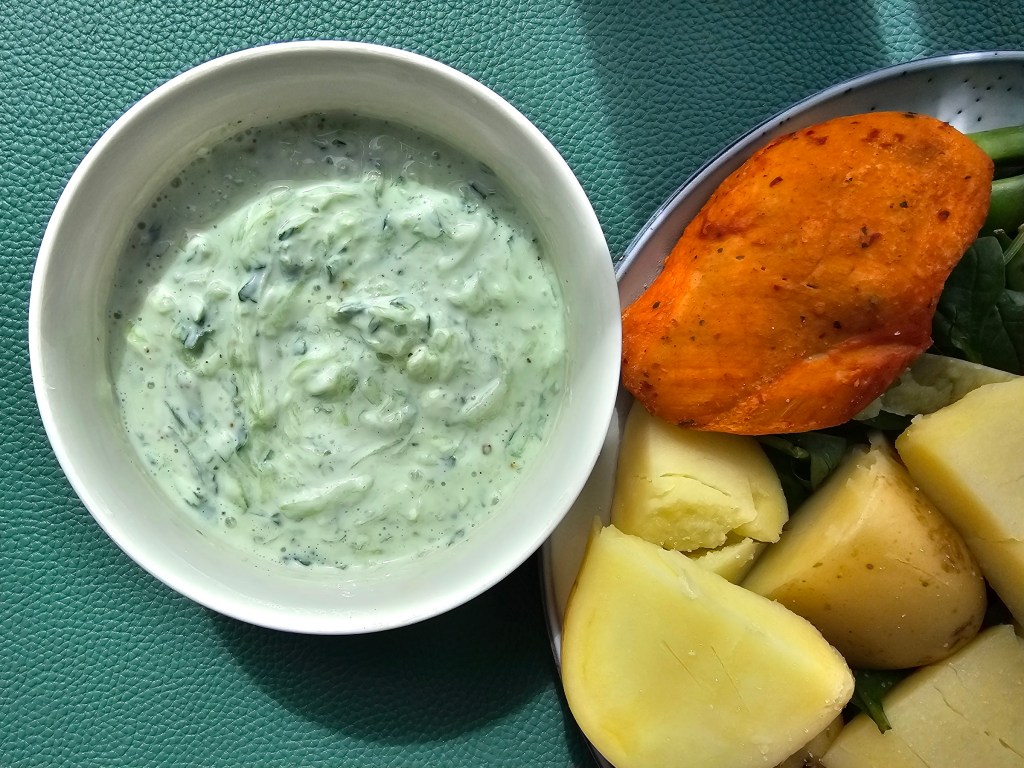 A bowl of Mizeria next to a plate with grilled chicken and potatoes.