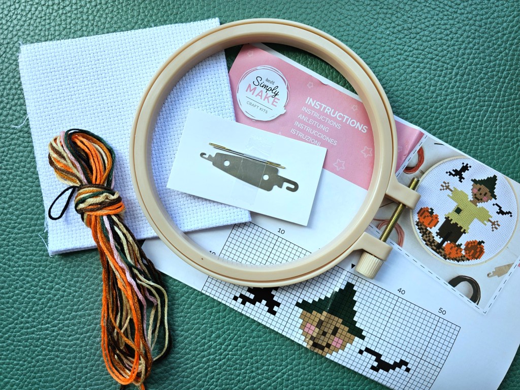 Contents of the DIY cross stitch kit with a Halloween scarecrow design. The kit included a blank white aida, a plastic hoop, two needles, a threader, yarn in assorted colours and a leaflet with the diagram and instructions.