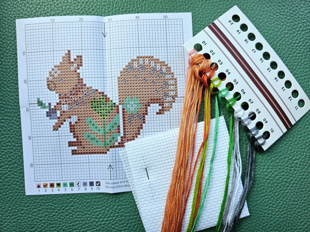Contents of the DIY cross stitch kit with a squirrel design - blank white aida, a sewing needle, yarn in 10 assorted colours and a leaflet with the pattern.
