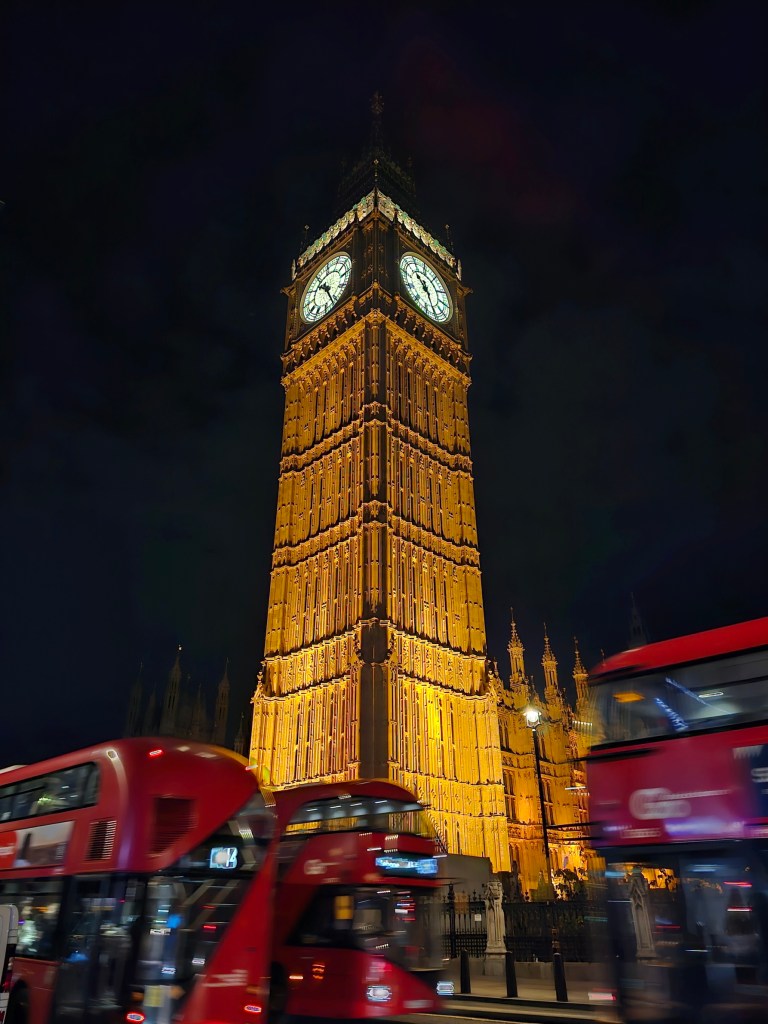 Illuminated Big Ben at night. Three red double-decker buses running past the Big Ben.