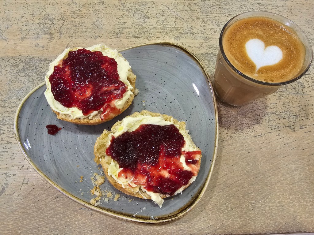 A scone sliced in half, with clotted cream and strawberry jam on each half. Served with a small cup of coffee.