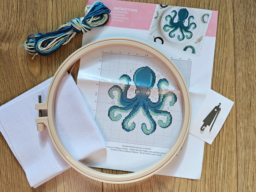 The contents of the pre assorted cross stitch kit, including a plastic hoop, blank aida, yarn in various shades of blue, beige and green, two needles, a threader and a leaflet with instructions.