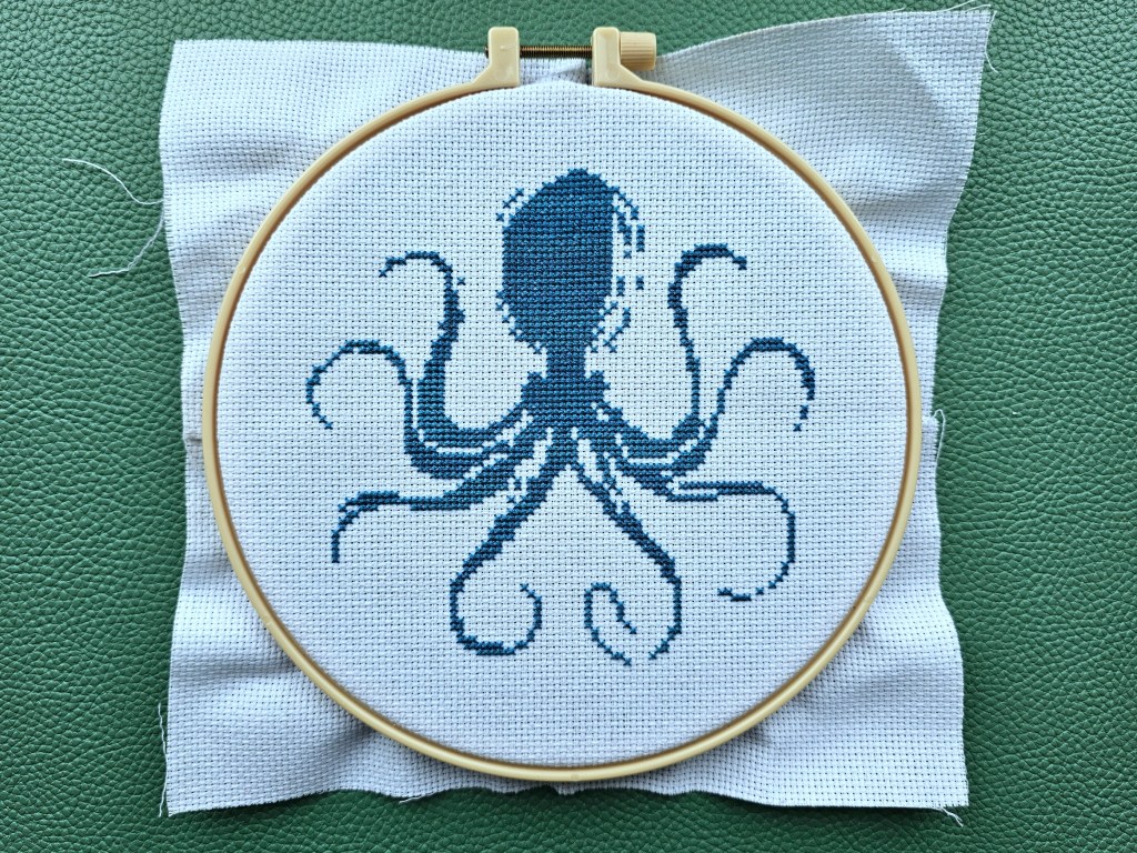 Blue octopus cross stitch design, partially completed. The most dominant shade of blue parts of the octopus's body is finished.