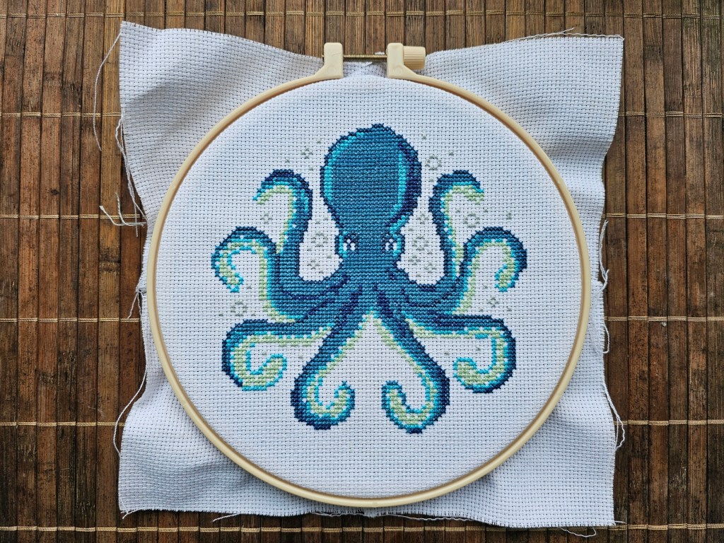 Blue octopus cross stitch design, partially completed. The blue and green parts of the octopus's body are finished, as well as grey bubbles around the animal.