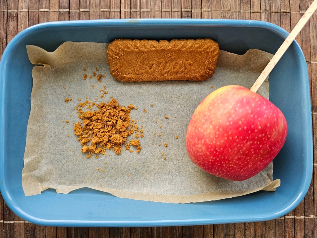 An apple on a stick next to a pile of crushed Biscoff biscuit.