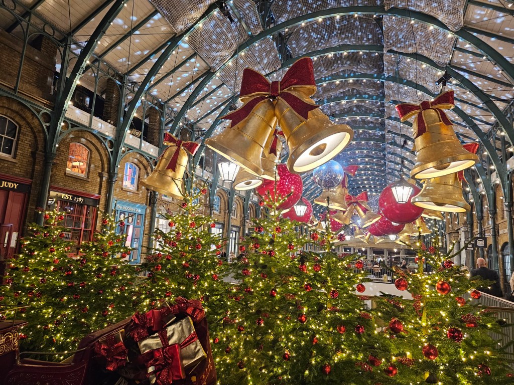 Christmas decorations in Covent Garden, London, in 2024. There are large bells with red bows under the roof alongside large, red baubles, and Christmas trees on the ground.
