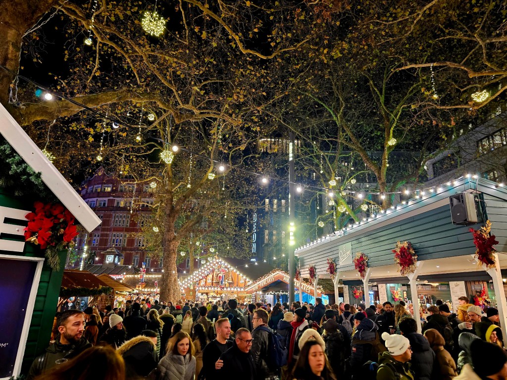 Christmas Market at Leicester Square, London. There are vendor stalls, fairly lights, and a large crowd.