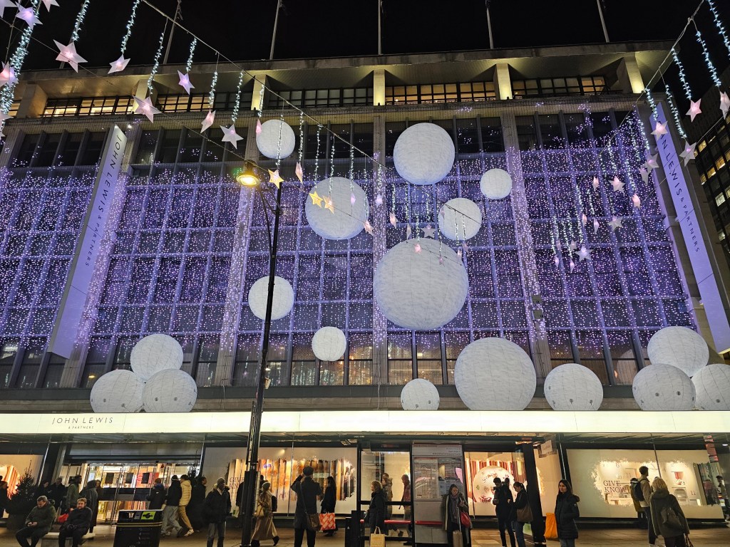 Christmas decorations outside of John Lewis store at Oxford Street, London. There are thousands of purple fairy lights and large, white snowballs.
