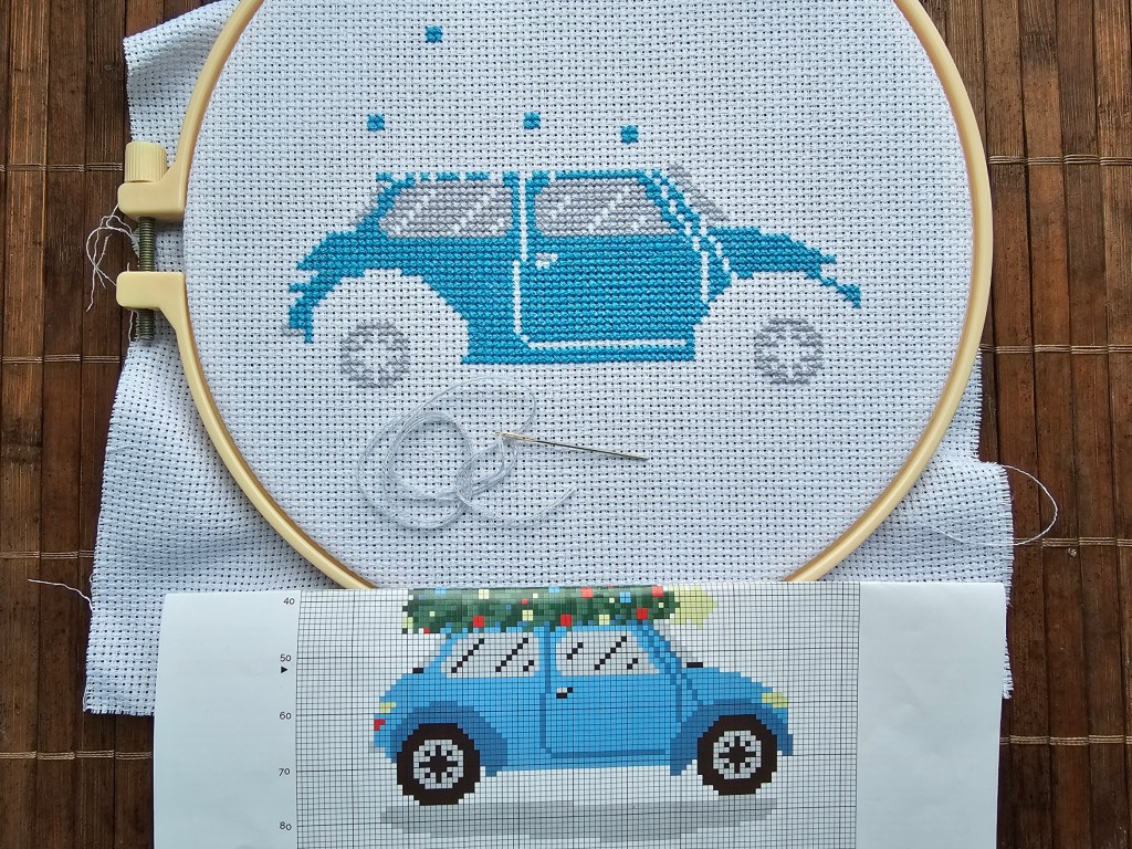 Partially finished cross stitch project depicting a car transporting a Christmas Tree. The blue parts are done, the grey parts are partially finished.