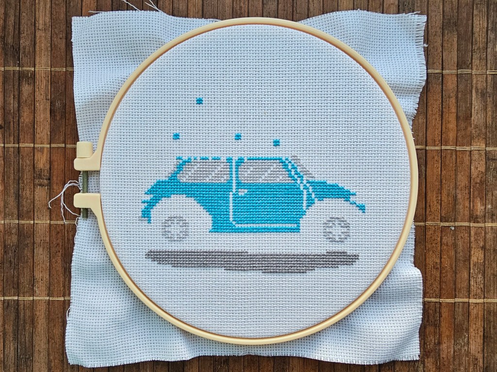 Partially finished cross stitch project featuring a blue car carrying a decorated Christmas tree.