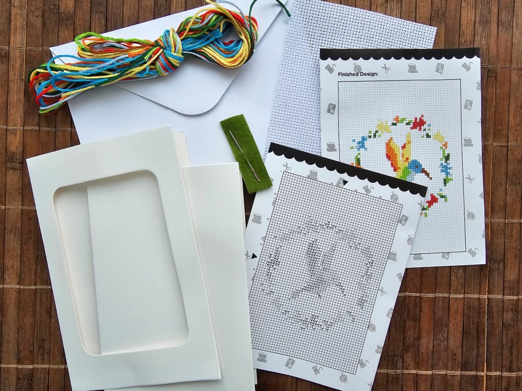 Contents of a cross stitch card making kit featuring a colourful hummingbird design - assorted yarn, instructions, aida and a card blank.