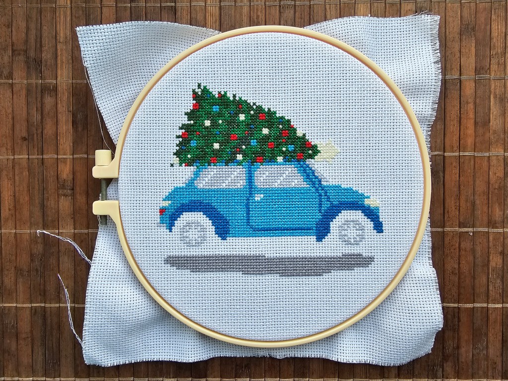 Nearly finished cross stitch project featuring a blue car carrying a decorated Christmas tree. Only black details are missing.