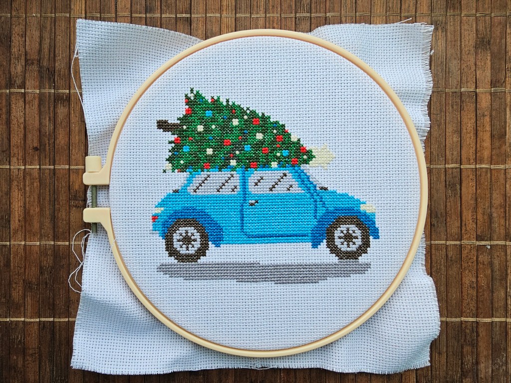 Finished cross stitch project featuring a blue car carrying a decorated Christmas tree.