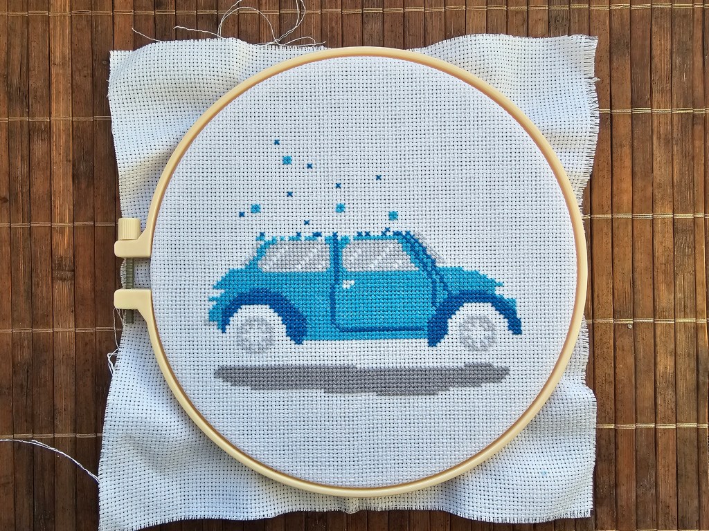 Partially finished cross stitch project featuring a blue car carrying a decorated Christmas tree.