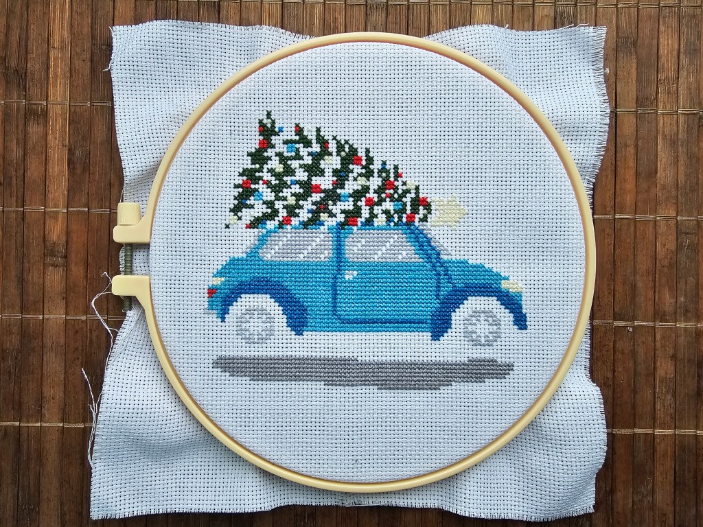 Nearly finished cross stitch project featuring a blue car carrying a decorated Christmas tree.