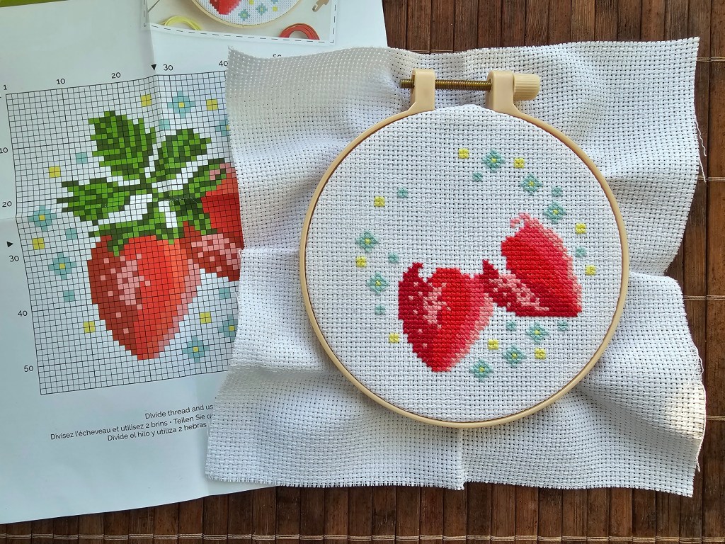 A plastic embroidery hoop with a white aida. All shades of red from a strawberry design are sewn, as well as small blue and yellow flowers.