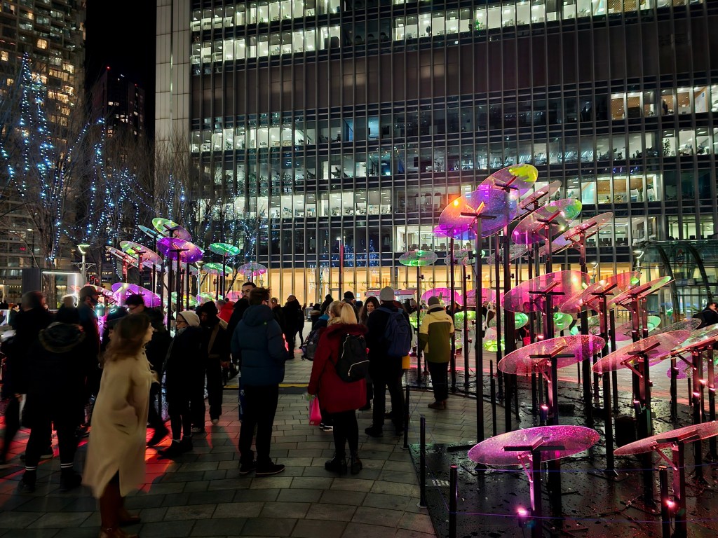 An abstract installation looking like a giant percussion illuminated with holographic lights. There's a crows of people admiring the exhibit.